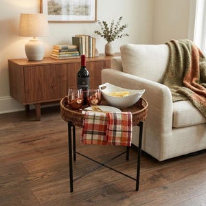 May include: A round brown woven wicker tray table with black metal legs, holding a bottle of red wine, two copper-toned glasses, a white bowl with snacks, a white plate with gold forks, and a folded red, orange, and white plaid cloth napkin.