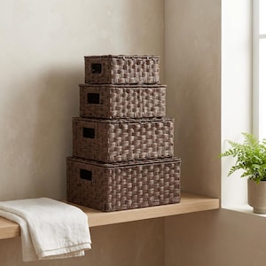 May include: Four brown woven storage boxes of varying sizes are stacked on a wooden shelf. Each box has a rectangular shape with a lid and cut-out handles. A white folded towel rests on the shelf below the boxes. A potted plant is visible in the background.