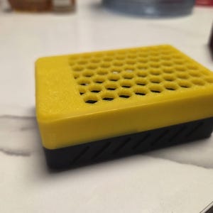 May include: A yellow and black rectangular device. The top is yellow with a honeycomb pattern of holes. The bottom is black with diagonal grooves. The device is sitting on a white surface.