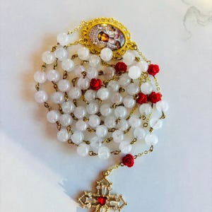 Könnte beinhalten: Ein Rosenkranz mit weißen Perlen, einer goldfarbenen Kette und roten Rosenakzenten. Der Rosenkranz hat ein goldfarbenes Kreuz mit einem roten Mittelstein und einen goldfarbenen ovalen Anhänger mit einem religiösen Bild.