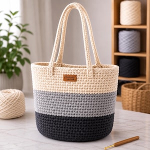 May include: A crochet tote bag featuring a color-blocked design with horizontal stripes in cream, light gray, and dark charcoal gray. The bag has two matching cream handles and a small brown rectangular leather tag labeled Handmade with Love.