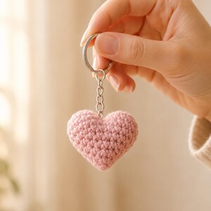 May include: A soft, dusty pink crocheted heart keychain featuring a textured yarn surface. The heart is attached to a silver-toned metal split ring by a short, four-link silver chain, creating a simple and functional accessory for keys or bags.