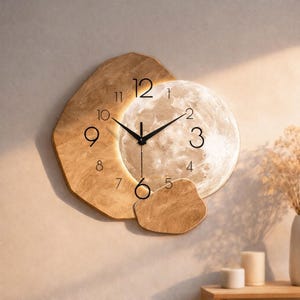 Pode incluir: Um relógio de parede decorativo com um design de lua. O mostrador do relógio é uma lua brilhante, rodeada por uma moldura de madeira. Os ponteiros do relógio são pretos e os números são indicados por linhas pretas simples. O relógio está montado numa parede de cor clara.