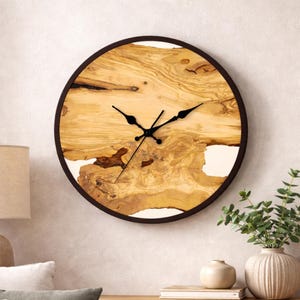 Puede incluir: Un reloj de pared redondo con una esfera de veta de madera natural y manecillas negras. La esfera del reloj está encerrada en un marco marrón oscuro. La veta de la madera tiene tonos marrones claros y oscuros con acentos blancos. El reloj está montado en una pared clara.