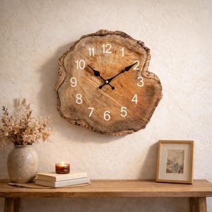 Puede incluir: Un reloj de pared rústico hecho de una sección transversal de tronco de árbol. La esfera del reloj tiene números blancos y manecillas negras. La veta natural de la madera y la forma irregular le dan una estética única. Diámetro aproximado de 30 cm.