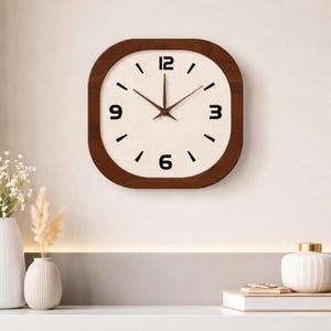 Puede incluir: Reloj de pared cuadrado de madera con esfera color crema y números negros. El reloj tiene un marco y manecillas de madera marrón oscuro. El reloj está montado en una pared de color claro, con jarrones decorativos y libros en primer plano.