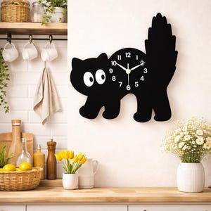 Könnte beinhalten: Eine schwarze, katzenförmige Wanduhr mit weißen Augen und einem Schwanz. Das Zifferblatt hat schwarze Zahlen und Zeiger. Die Uhr ist an einer weißen Wand in einer Küchenumgebung angebracht.