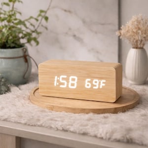 Puede incluir: Un reloj digital rectangular de madera que muestra la hora 1:58 y la temperatura 20°C. El reloj está sobre una bandeja de madera sobre una superficie blanca y esponjosa. Una pequeña planta y un jarrón están en el fondo.