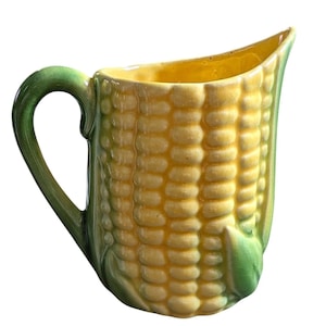 May include: A ceramic pitcher shaped like a corn cob with a yellow kernel body and green husk accents. The handle is formed from a green leaf design, and the interior of the vessel is a smooth, glossy yellow finish with a visible crackle glaze.
