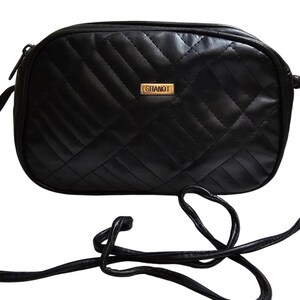 May include: A black crossbody bag featuring a quilted pattern with diagonal stitching. The bag has a rectangular shape with rounded corners and a gold-toned metal plaque on the front center with the brand name GITANO printed in black text.