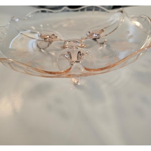 May include: A vintage, pink glass serving dish with a scalloped edge and three delicate feet. The dish features etched floral designs and a central support. The glass has a subtle, translucent quality, and the dish is likely used for serving desserts or small treats.