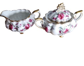 Juego de cremera y azucarero de porcelana rosa vintage con base dorada y diseño floral.