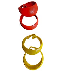 May include: Two pairs of hoop earrings in bright colors. One pair is a vibrant red, and the other is a sunny yellow. The earrings have a rounded, chunky design with a clip closure. The earrings are made of a glossy material.