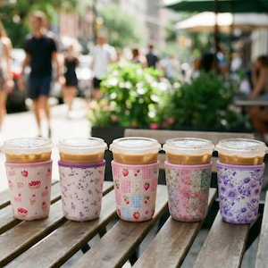 Könnte beinhalten: Fünf Eiskaffeebecher mit durchsichtigen Kunststoffdeckeln, jeweils in einer Stoffhülle. Die Hüllen zeigen verschiedene Muster, darunter Erdbeeren, kleine violette Blumen, ein rosa-blau kariertes Erdbeermuster und florale Designs.
