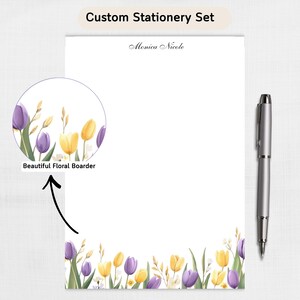 Peut inclure: Ensemble de papeterie personnalisé avec un bloc-notes blanc orné d'une bordure florale de tulipes violettes et jaunes. Un stylo argenté est posé à côté du bloc-notes. Le texte "Custom Stationery Set" et "Monica Nicole" sont visibles.