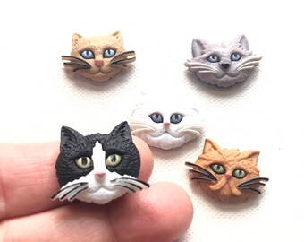 May include: A collection of five cat face embellishments in various colors. The cats have detailed features, including whiskers and eyes in shades of blue, green, and yellow. The cat faces are in shades of tan, gray, white, black, and orange.