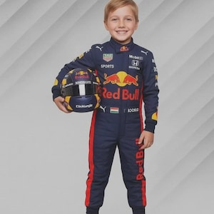 Könnte beinhalten: Ein Kind in einem marineblauen Red Bull Rennanzug mit roten Akzenten und passendem Helm. Der Anzug zeigt Sponsorenlogos und den Schriftzug "Red Bull". Der Helm ist marineblau, gelb und rot.