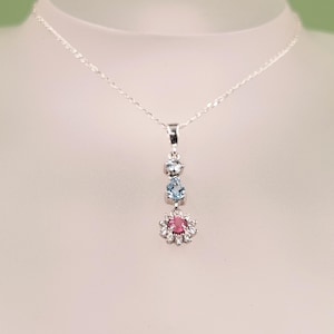 Puede incluir: Un collar de plata con un colgante vertical que presenta dos piedras azul claro, una redonda y otra en forma de pera, suspendidas sobre un dije en forma de flor. El dije contiene una piedra rosa central rodeada de pequeñas piedras blancas transparentes.