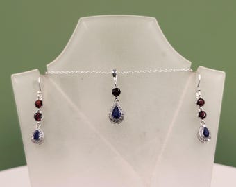 May include: A silver necklace and earring set displayed on a white mannequin. The necklace features a pendant with a dark red stone and a blue teardrop-shaped stone. The earrings have a similar design, with three dark red stones and a blue teardrop.