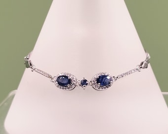 Op de afbeelding: Een zilveren armband met drie ovale, donkerblauwe edelstenen, elk omringd door kleinere, heldere stenen. De delicate ketting heeft een textuurpatroon. De sieraden worden gepresenteerd op een wit oppervlak.