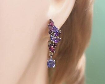 May include: A silver earring with a cascade of purple and violet gemstones. The earring features a square-cut, light purple stone at the bottom, with a cluster of teardrop-shaped stones in varying shades of purple and violet ascending upwards.