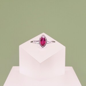 May include: A silver-toned ring featuring a central marquise-cut vibrant pink gemstone. The stone is surrounded by a halo of small, clear, sparkling accent stones set into the metal band, creating a refined and symmetrical jewellery piece.