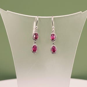 Puede incluir: Pendientes colgantes plateados con piedras rojas ovaladas, cada una rodeada de pequeñas piedras claras. Las piedras rojas miden aproximadamente 0,8 cm de largo.