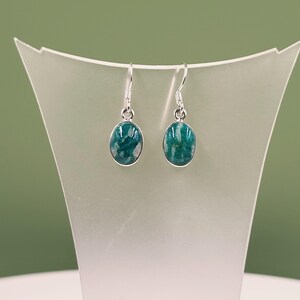 Peut inclure: Boucles d'oreilles pendantes en argent avec des pierres ovales de couleur teal. Les pierres présentent un aspect marbré. Les boucles d'oreilles sont présentées sur un présentoir blanc sur fond vert. Bijoux.