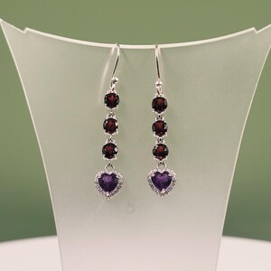 May include: A pair of silver drop earrings featuring three round, deep red garnet stones stacked vertically above a single heart-shaped purple amethyst stone, which is surrounded by a halo of small, clear, sparkling white accent stones.
