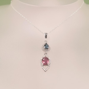 May include: A silver-toned necklace featuring a pendant with two stacked, marquise-shaped frames lined with small clear stones. The top frame holds a small, dark blue teardrop-shaped stone, while the bottom frame holds a larger, oval-shaped pink stone.