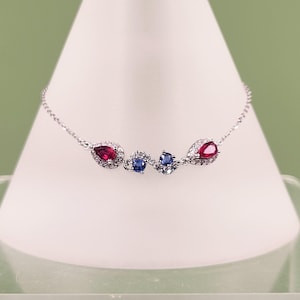 May include: A silver bracelet with a series of gemstones. The bracelet features two pear-shaped red gemstones, each surrounded by small clear stones. Between them are two blue gemstones and small clear stones.
