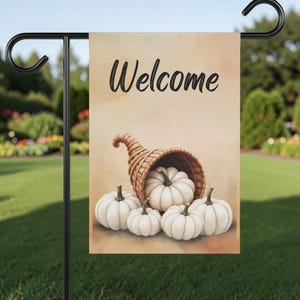 May include: A rectangular garden flag hanging on a black metal stand. The flag features a light tan background with the word Welcome in black script. A woven cornucopia basket sits at the bottom, spilling out five white pumpkins.