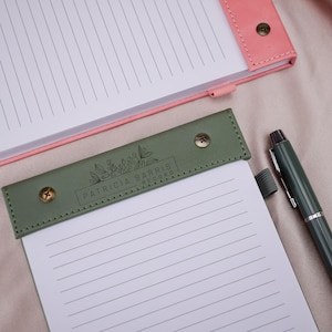 Può includere: Primo piano di un blocco note e di una penna. Il blocco note ha una copertina rosa e un risvolto in similpelle verde con la scritta "PATRICIA BARRIS RECORD". Una penna verde con la punta argentata è accanto al blocco note.