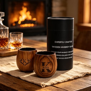 May include: Two rounded wooden whiskey glasses with dark brown grain patterns, each featuring a laser-engraved letter K. One glass includes the name Kelsey. They are displayed next to a black cylindrical gift tube labelled EXPERTLY CRAFTED.