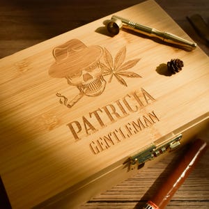 May include: A light-colored wooden box with a skull wearing a fedora, smoking a cigar, and a cannabis leaf etched on the lid. The name "PATRICIA" and the word "GENTLEMAN" are also etched on the box.