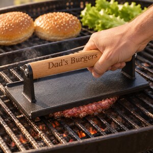 May include: A rectangular black cast iron burger press with a light brown wooden handle featuring the engraved text "Dad's Burgers". The tool is positioned on a metal grill grate, pressing down on a raw meat patty with burger buns nearby.