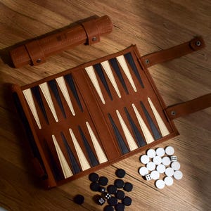 Pode incluir: Um conjunto de gamão castanho com um estojo enrolado, um tabuleiro aberto e peças de jogo. O tabuleiro apresenta triângulos pretos, brancos e bege alternados. Peças de jogo e dados pretos e brancos estão espalhados ao redor do tabuleiro.