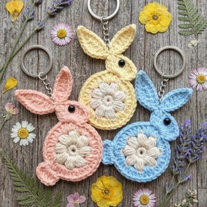May include: Three handmade crochet bunny keychains in pastel colors: pink, yellow, and blue. Each bunny has a flower design in the center and a black bead for the eye. The keychains are on silver rings, and the background is a wooden surface with dried flowers.