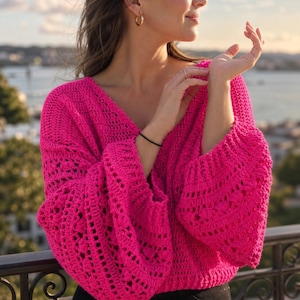 May include: A bright pink, open-weave crochet sweater with a deep V-neck and long, flared sleeves. The sweater is made of a soft, textured yarn and has a relaxed fit. The background shows a body of water and a cityscape.