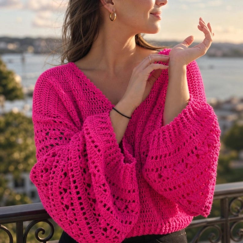 May include: A bright pink, open-weave crochet sweater with a deep V-neck and long, flared sleeves. The sweater is made of a soft, textured yarn and has a relaxed fit. The background shows a body of water and a cityscape.