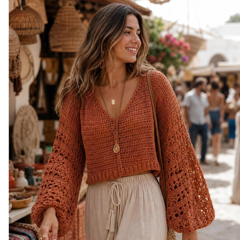May include: A rust-colored crochet top with a deep V-neck and long, bell sleeves. The top is paired with light beige pants. The image is taken outdoors, with a blurred background of a market setting.