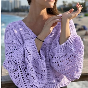 May include: A light purple crochet sweater with a V-neck and long, flared sleeves. The sweater features an openwork design with a textured pattern. The sleeves are gathered at the wrist. The sweater is made of a soft, lightweight material.