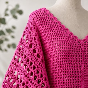 May include: A vibrant fuchsia crochet top with a V-neckline and scalloped edges. The sleeves feature openwork detailing, adding a unique texture to the garment. The top is displayed on a mannequin.