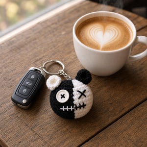 May include: A split-color black and white crocheted bear head keychain featuring a white button eye, a stitched X eye, and a stitched mouth. The keychain is attached to a silver metal ring connected to a black plastic car key fob.