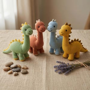 Può includere: Quattro giocattoli di dinosauri all'uncinetto nei toni del verde, corallo, blu e giallo. Ogni dinosauro ha un collo lungo, zampe piccole e una fila di punte sulla schiena. I giocattoli sono disposti su una superficie neutra con pietre e lavanda.