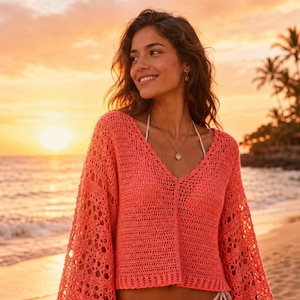 May include: Coral-colored crochet sweater with a V-neck and long, flared sleeves. The open-weave design allows glimpses of the skin beneath. The sweater is worn on a beach at sunset.