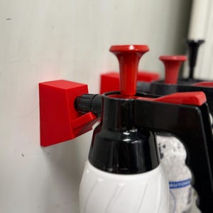 Op de afbeelding: Rode en zwarte spuitflessen met witte etiketten zijn aan een muur bevestigd. De flessen hebben zwarte handvatten en rode pompen. De etiketten lezen "INDUSTRIAL AUTOMOTIVE". Een rode beugel houdt de flessen vast.