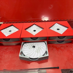 Könnte beinhalten: Rote Aufbewahrungsboxen mit rautenförmigen Aussparungen, die graue Schleifscheiben enthalten. Die Boxen sind mit Zahlen wie 320, 400 und 600 beschriftet, die die Körnungen angeben. Das Set befindet sich auf einer roten Oberfläche.