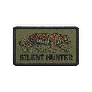 May include: Embroidered patch featuring a tiger walking through tall grass. The tiger has orange and black stripes. The patch is rectangular with a black border and the words "SILENT HUNTER" in dark letters.