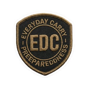 May include: A black and tan patch with the text "EVERYDAY CARRY - PREPAREDNESS" encircling a central "EDC" design. The patch has a shield shape with a tan border and white backing.